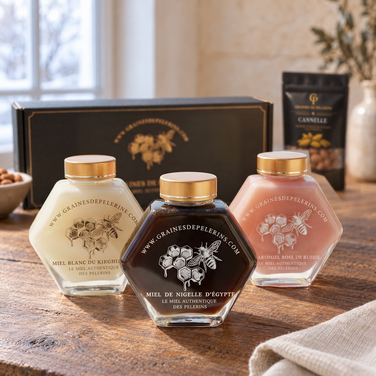 Coffret Découverte : miel blanc, miel de Nigelle et Aromiel Rose, devant leur grand écrin. Amandes Cannelle en arrière-plan flou.