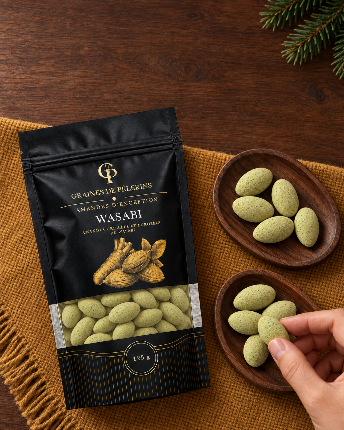 Une main prend une amande Wasabi dans une coupelle, près du sachet sur un tissu ocre.